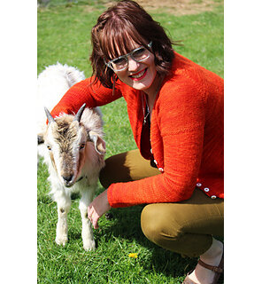 image is of female wearing an orange cardigan crouched down with arm around goat