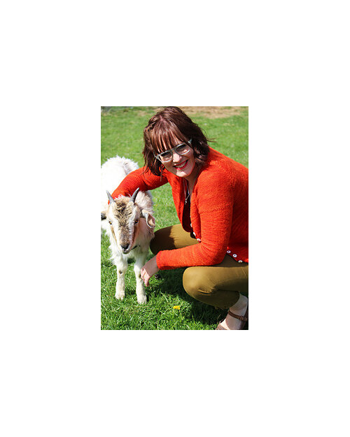 image is of female wearing an orange cardigan crouched down with arm around goat