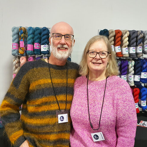 image shows a blonde haired lady and a bearded man standing behind a yarn stall