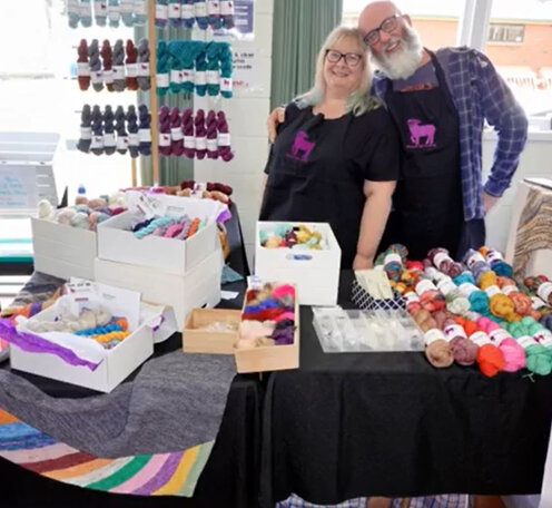 image shows a blonde haired lady and a bearded man standing behind a yarn stall