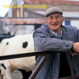 Rural farmer leans on fence with cow and cow shed behind him